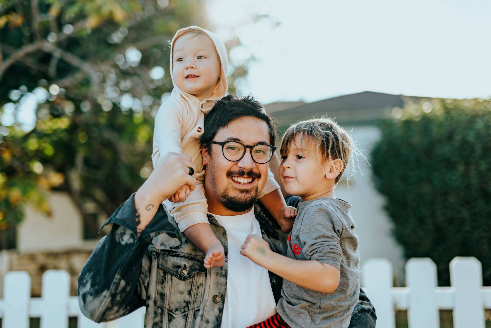 man carrying two children outside a suburban house with a white picket fence